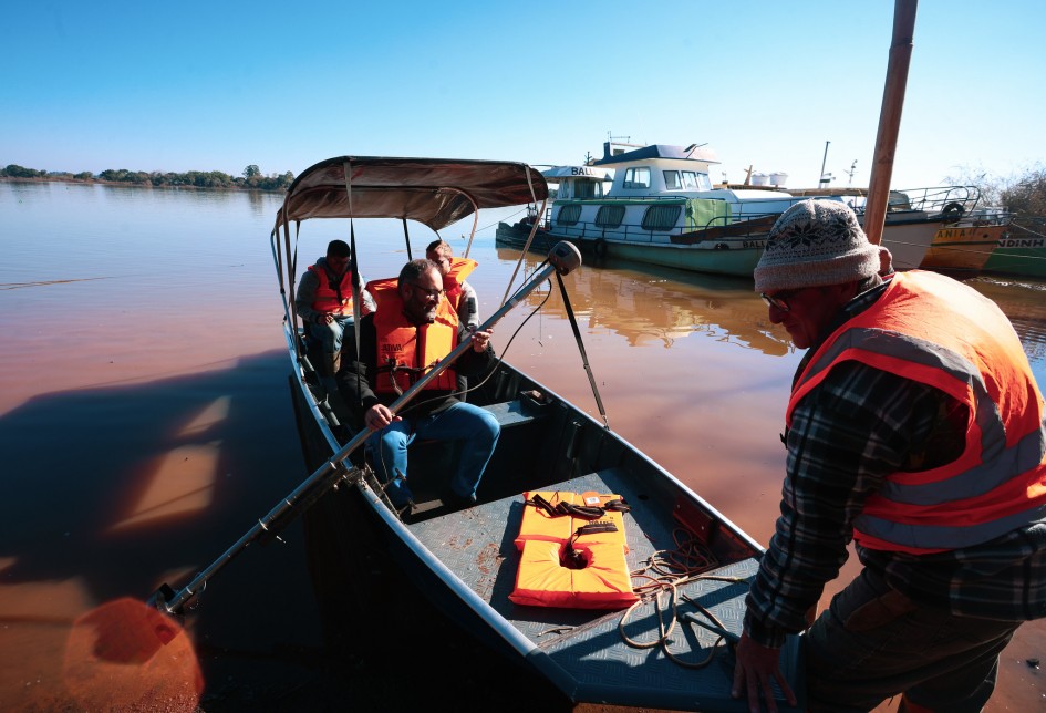 Uma fotografia em nível dos olhos mostra um barco de metal com cobertura de lona em um rio de água barrenta. Dentro do barco, três homens usando coletes salva-vidas laranjas observam um quarto homem, à direita, que está na beira do barco, usando um colete salva-vidas laranja sobre uma camisa xadrez e um gorro cinza. Um dos homens sentados no barco segura um remo. Ao fundo, outras embarcações estão ancoradas. O céu é azul e o sol é forte.