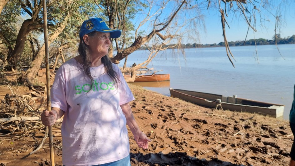Uma mulher idosa, de pele clara e cabelos castanhos longos, está em primeiro plano, olhando para a direita com uma expressão pensativa. Ela veste uma camiseta rosa com letras em verde-limão e um boné azul com um emblema. Em sua mão esquerda, ela segura um longo galho de madeira. Ela está em uma margem de rio marrom-avermelhada, com o solo úmido e irregulares. Atrás dela, várias árvores com galhos secos se estendem sobre a água. No rio, que tem uma tonalidade marrom-clara, há dois barcos pequenos, um laranja e outro cinza, próximos à margem. Ao fundo, o rio se estende sob um céu azul claro, sem nuvens.