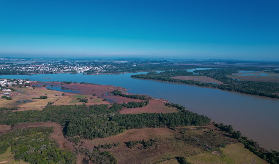 Um panorama aéreo mostra um rio largo e sinuoso que corta uma paisagem verde e marrom sob um céu azul claro. No canto superior esquerdo, uma cidade com construções claras e telhados escuros se alonga junto à margem do rio. A seguir à cidade, o rio se expande em áreas de águas mais escuras, com pequenas ilhas de terra e vegetação em seu leito. A paisagem é formada por campos agrícolas marrons e verdes, misturados com trechos de mata fechada e escura. Ao longe, observa-se uma planície sob uma suave névoa azul no horizonte.