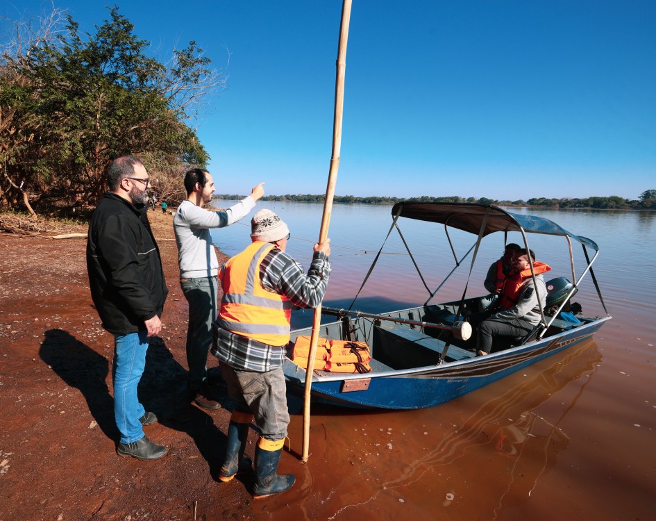 A imagem mostra cinco pessoas próximas a um rio. Três estão em pé na margem, enquanto duas estão sentadas em um pequeno barco, vestindo um colete salva-vidas laranja. Uma das pessoas na margem segura um bastão longo, possivelmente para ajudar a atracar ou empurrar o barco. Ao fundo, há árvores e céu azul claro.